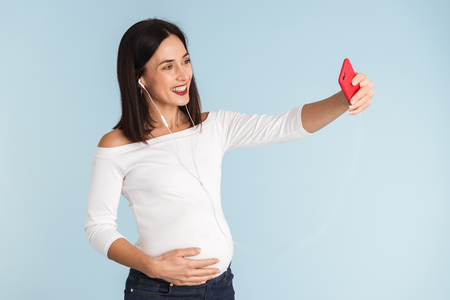 Photo of a young pregnant woman isolated over blue wall background take a selfie by mobile phone.の写真素材