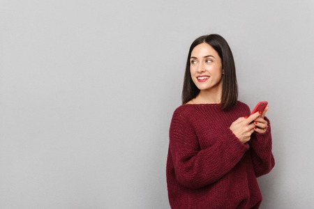 Picture of pretty young woman dressed in burgundy sweater using mobile phone isolated over grey wall background.の写真素材