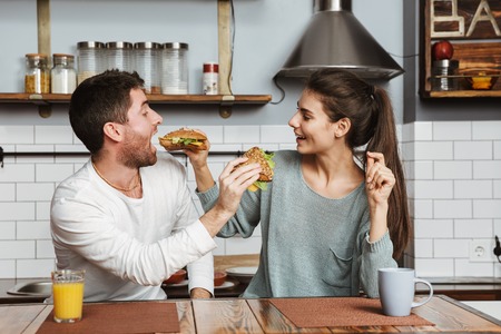 Young couple sitting at the kitchen during breakfast at home, feeding each other with sandwichesの写真素材