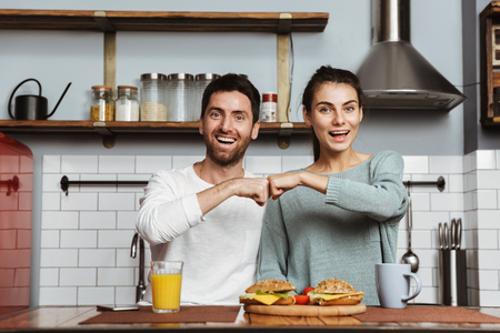Happy young couple sitting at the kitchen during breakfast at home, fist bumpの写真素材