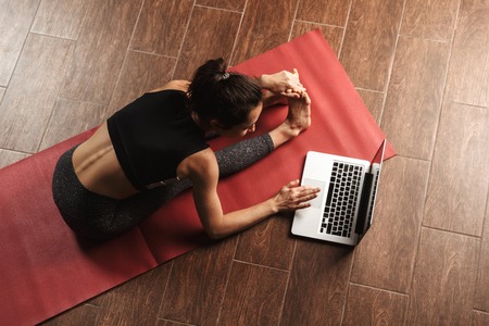 Beautiful healthy woman doing yoga exercises while sitting on a fitness mat at home, using laptop computer, stretchingの写真素材