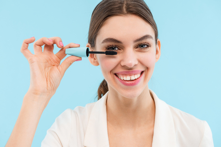 Photo of beautiful young woman posing isolated over blue background holding lash mascara doing makeup.の写真素材