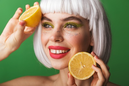 Attractive young topless woman wearing short white hair standing isolated over green background, showing sliced lemonの写真素材
