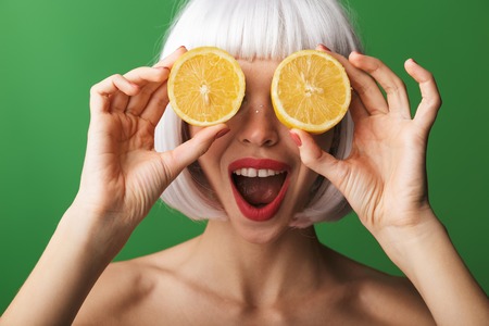 Attractive young topless woman wearing short white hair standing isolated over green background, showing sliced lemonの写真素材