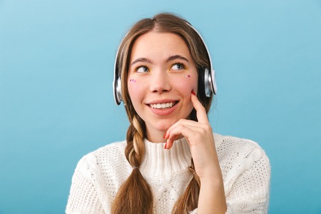 Lovely young girl wearing winter clothes standing isolated over blue background, listening to music...