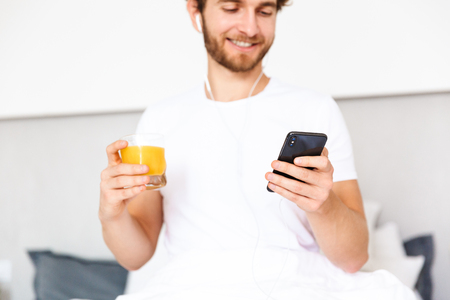 Image of a handsome young bearded man at home listening music with earphones using mobile phone drinking juice.の写真素材