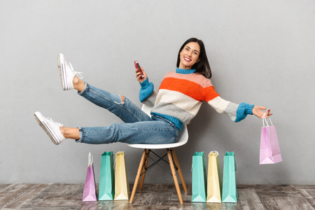 Image of attractive woman 30s sitting in chair with colorful shopping bags and using mobile phone isolated over gray backgroundの写真素材