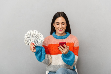Photo of positive woman 30s holding bunch of money and smartphone isolated over gray backgroundの写真素材