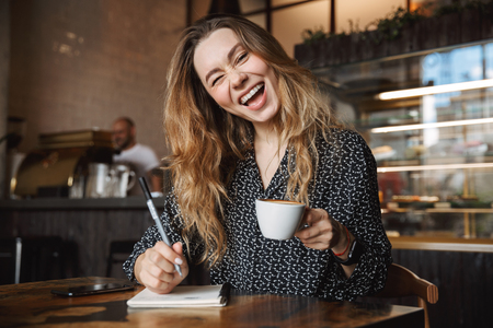Photo of beautiful young pretty woman sitting in cafe indoors writing notes in notebook drinking coffee winking.の写真素材