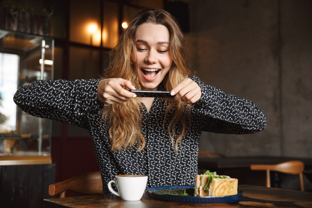 Photo of an emotional young pretty woman sitting in cafe indoors take a photo of her breakfast by mobile phone.の写真素材