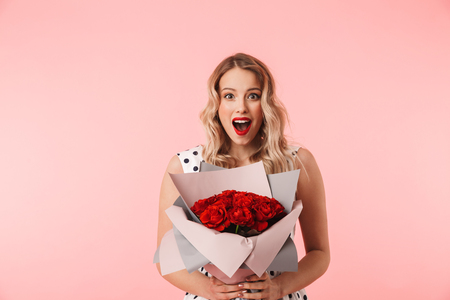 Beautiful young blonde woman wearing dress standing isolated over pink background, holding bouquet of rosesの写真素材