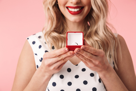 Close up of a beautiful young blonde woman wearing dress standing isolated over pink background, holding box with an engagement ring insideの写真素材