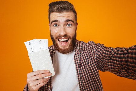 Portrait of a cheerful young man wearing casual clothes standing isolated taking a selfie, showing passport with flight ticketsの写真素材