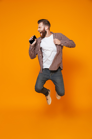 Full length portrait of a cheerful young man wearing casual clothes isolated jumping, listening to music with earphones, holding mobile phoneの写真素材