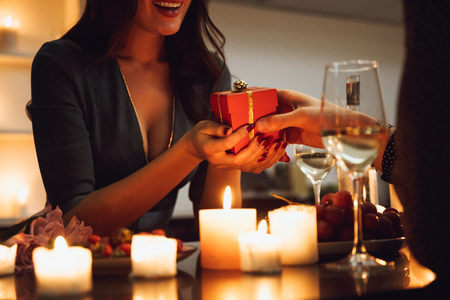 Beautiful passionate couple having a romantic candlelight dinner at home, drinking wine, woman getting a gift boxの写真素材