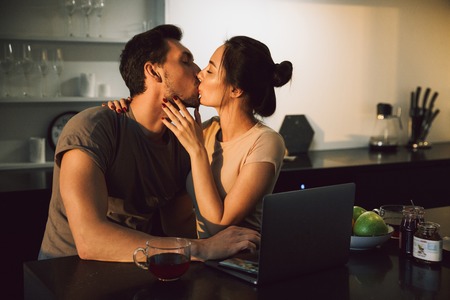 Beautiful young couple embracing while sitting at the kitchen table with laptop computer