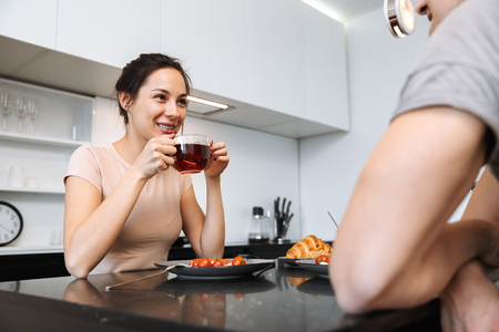 Image of a young happy loving couple indoors in home at kitchen have a breakfast drinking tea.の写真素材