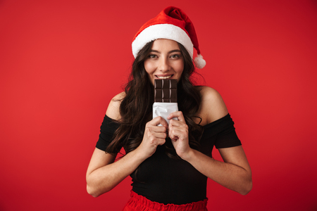 Cheerful young woman wearing Christmas hat standing isolated over red background, eating chocolateの写真素材