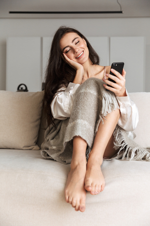 Photo of smiling woman 20s wrapped in blanket sitting on sofa at home and using smartphoneの写真素材
