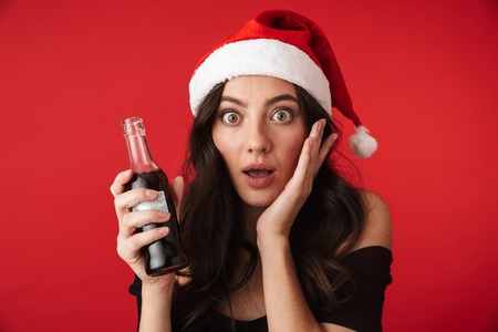 Cheerful young woman wearing Christmas hat standing isolated over red background, showing glass bottle with fizzy drinkの写真素材