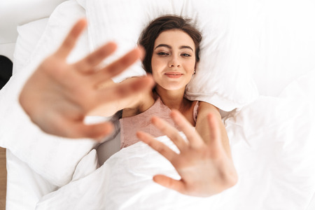 Photo from above of brunette woman 20s smiling while lying in bed on white pillow after sleep or napの写真素材