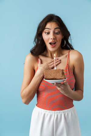Lovely cheerful girl wearing summer clothes standing isolated over blue background, eating birthday cakeの写真素材