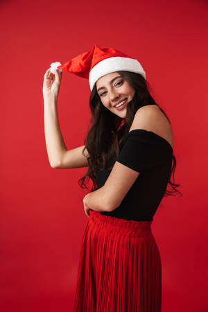 Excited young woman wearing Christmas hat standing isolated over red background, posingの写真素材