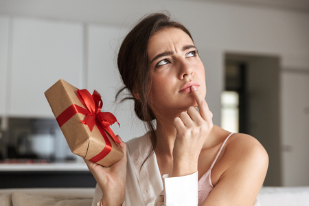 Portrait of adorable woman 20s in leisure wear sitting on couch in cozy apartment and holding present boxの写真素材