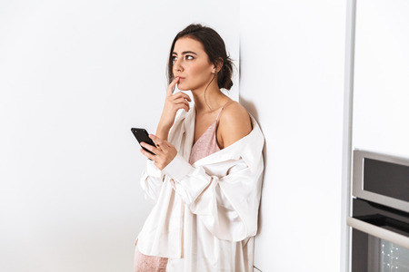 Photo of pretty woman 20s using earphones while standing in kitchen at homeの写真素材