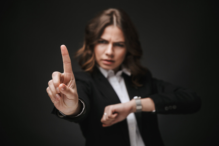 Portrait of a confident young businesswoman wearing formal suit standing isolated over black background, checking timeの写真素材