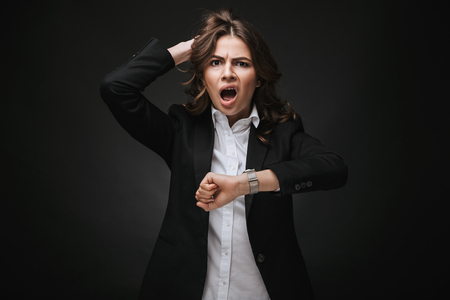 Shocked young businesswoman wearing a suit standing isolated over black background, checking timeの写真素材