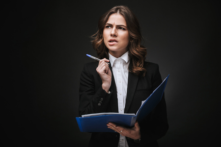 Portrait of a tired busy young businesswoman wearing formal suit standing isolated over black background, holding folder with documentsの写真素材