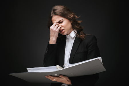 Portrait of a tired busy young businesswoman wearing formal suit standing isolated over black background, holding folder with documentsの写真素材
