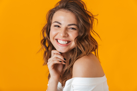 Portrait of a cheerful young woman wearing white shirt standing isolated over yellow background, looking at camera, posingの写真素材