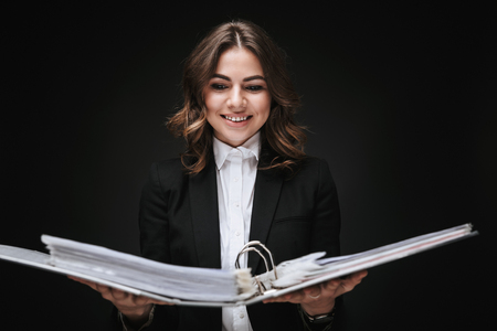 Portrait of a confident young businesswoman wearing formal suit standing isolated over black background, holding folder with documentsの写真素材