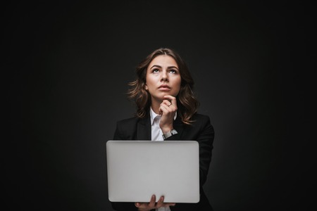 Confident young businesswoman wearing a suit standing isolated over black background, holding laptop computer, looking upの写真素材