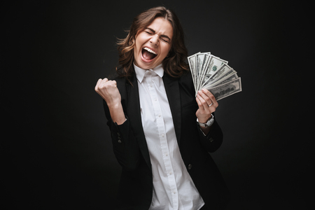 Portrait of a confident young businesswoman wearing formal suit standing isolated over black background, holding bunch of money banknotesの写真素材