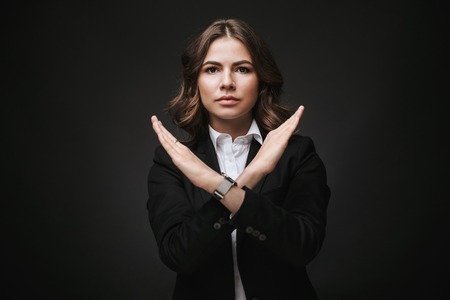 Portrait of a serious confident young businesswoman wearing formal suit standing isolated over black background, showing stop gestureの写真素材