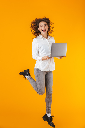 Full length portrait of a cheerful young woman wearing white shirt jumping isolated over yellow background, holding laptopの写真素材