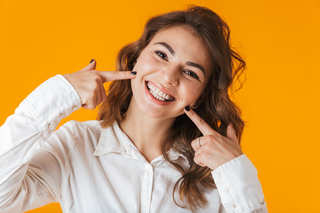 Portrait of a cheerful young woman wearing white shirt standing isolated over yellow background, looking at camera, posingの写真素材