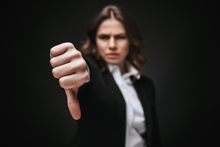 Portrait of a confident young businesswoman wearing formal suit standing isolated over black background, showing thumbs downの写真素材