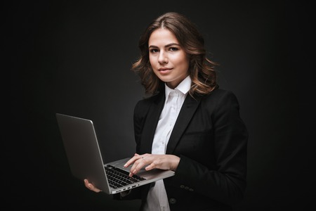 Portrait of a confident young businesswoman wearing formal suit standing isolated over black background, working on laptopの写真素材