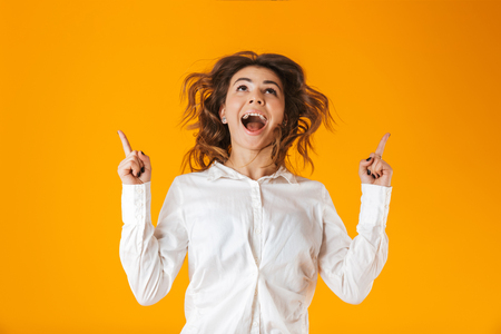 Portrait of a cheerful young woman wearing white shirt standing isolated over yellow background, pointing finger at copy spaceの写真素材
