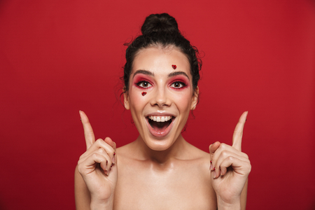 Image of a beautiful happy cheerful young woman with red bright makeup isolated over red wall background posing with hearts on face pointing to copyspace.の写真素材
