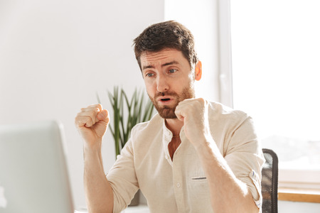 Photo of attractive businessman 30s wearing white shirt using laptop while working in bright office