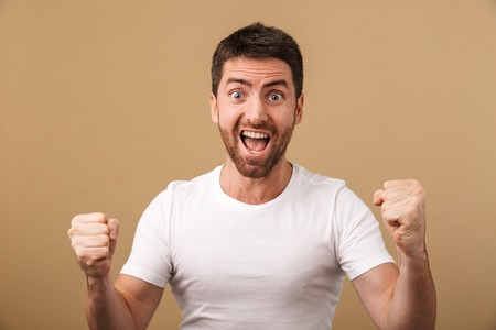Portrait of a cheerful young man standing isolated over beige background, celebrating successの写真素材