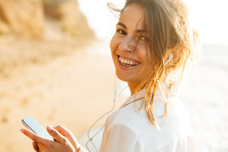 Image of gorgeous girl 20s smiling and using mobile phone while walking by seasideの写真素材