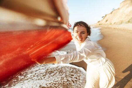 Portrait of young girl 20s smiling and walking with waving scarf along seashore
