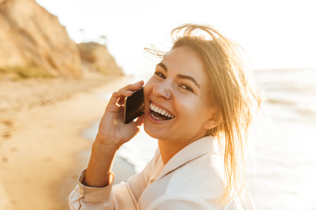 Image of european girl 20s smiling and talking on mobile phone while walking by seasideの写真素材