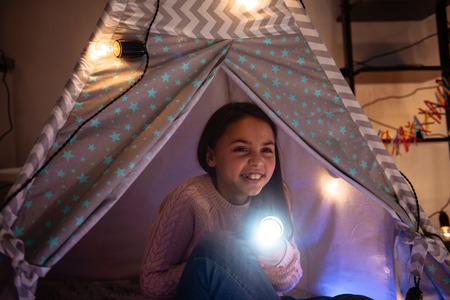 Cute little girl sitting inside childrens tent in a dark, holding flashlight. Christmas conceptの写真素材